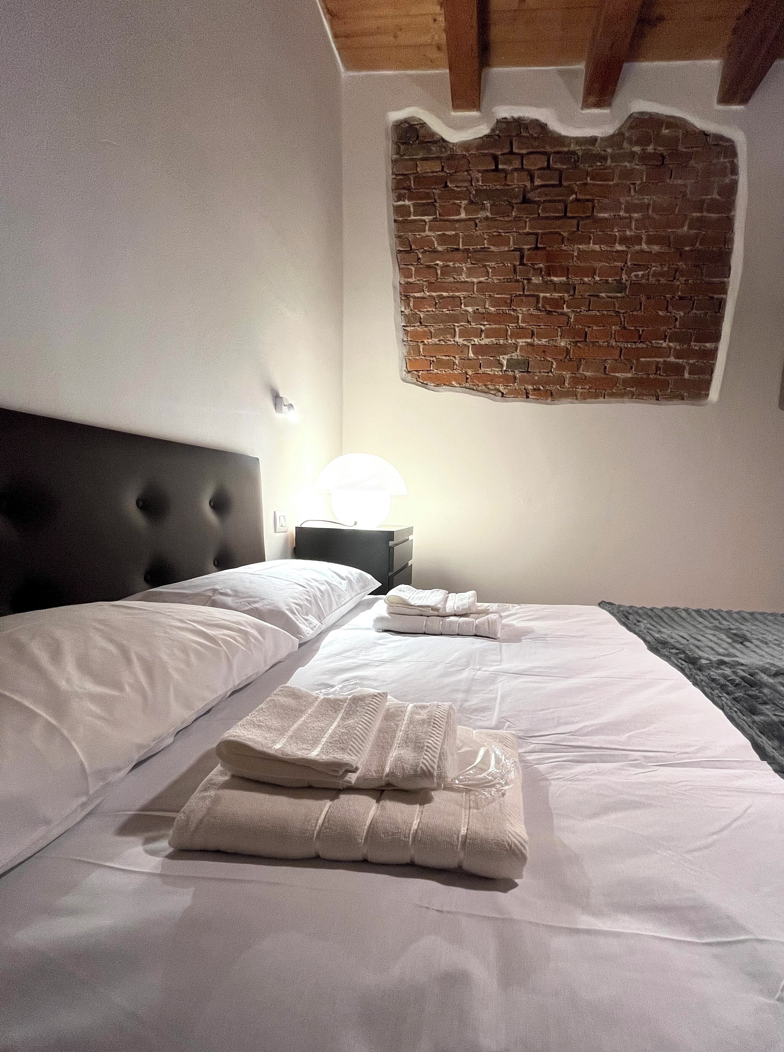 Une chambre d’hôtel avec un mur en briques apparentes, un lit soigneusement fait avec du linge de lit blanc et une tête de lit.
