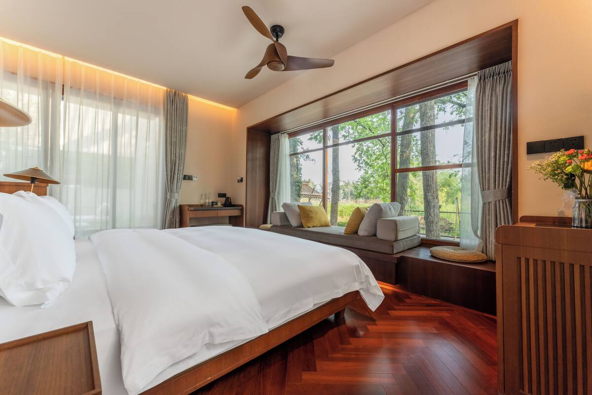 Ein Schlafzimmer mit einem großen Bett, einem Deckenventilator, einer Sitzbank unter dem Fenster mit Kissen und Blick auf Bäume.