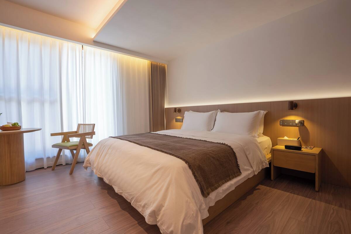 Ein modernes Hotelzimmer mit einem großen Bett, einem Nachttisch mit Lampe, einem Sessel und einem kleinen Tisch mit Obst.