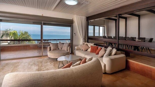 A spacious living area with a large window overlooking the sea, a sofa set, and a dining area with a high ceiling and modern design.