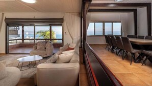 A spacious living area with a sofa set, a coffee table, and a dining table with chairs. Large windows provide a view of the outdoors.