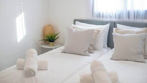 Un letto rifatto con lenzuola bianche e cuscini a righe, un comodino con una pianta e una finestra con tende leggere.