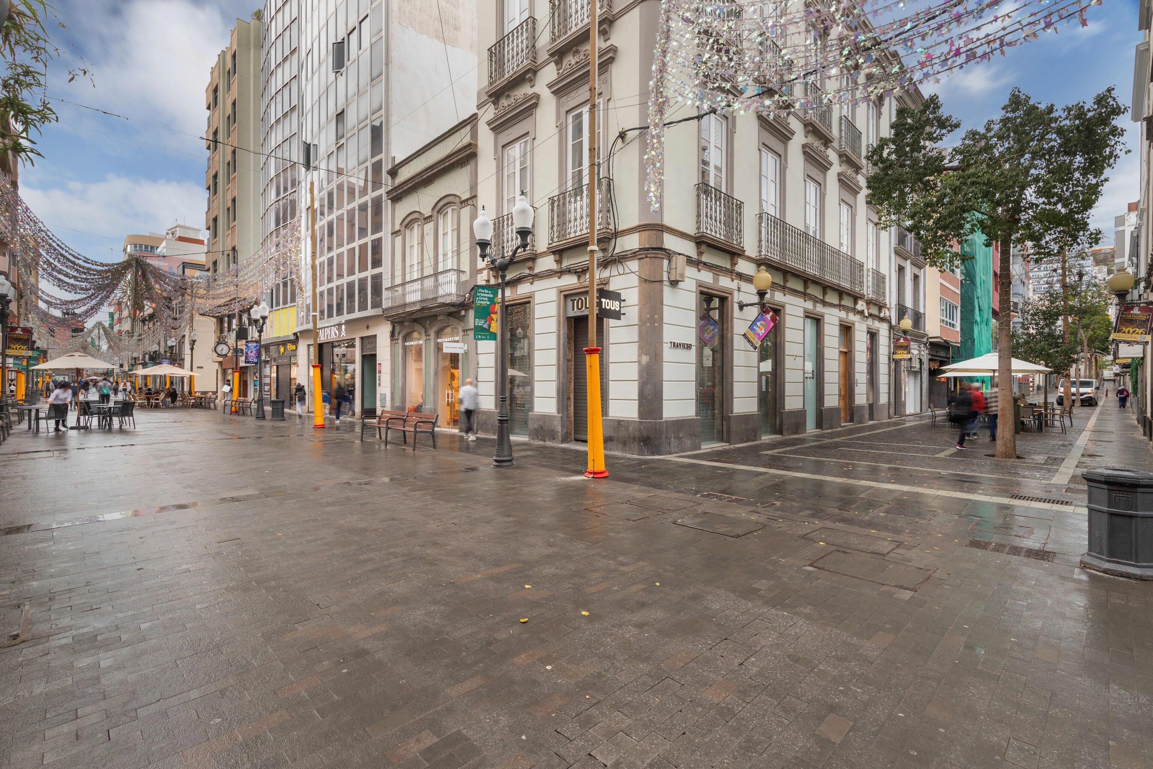 A street with buildings, a wet pavement, and a few people walking.