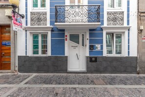 Un edificio con una puerta blanca identificada con 'TRAVIES C.M', un balcón con barandillas de hierro decorativas y una fachada azul y blanca.
