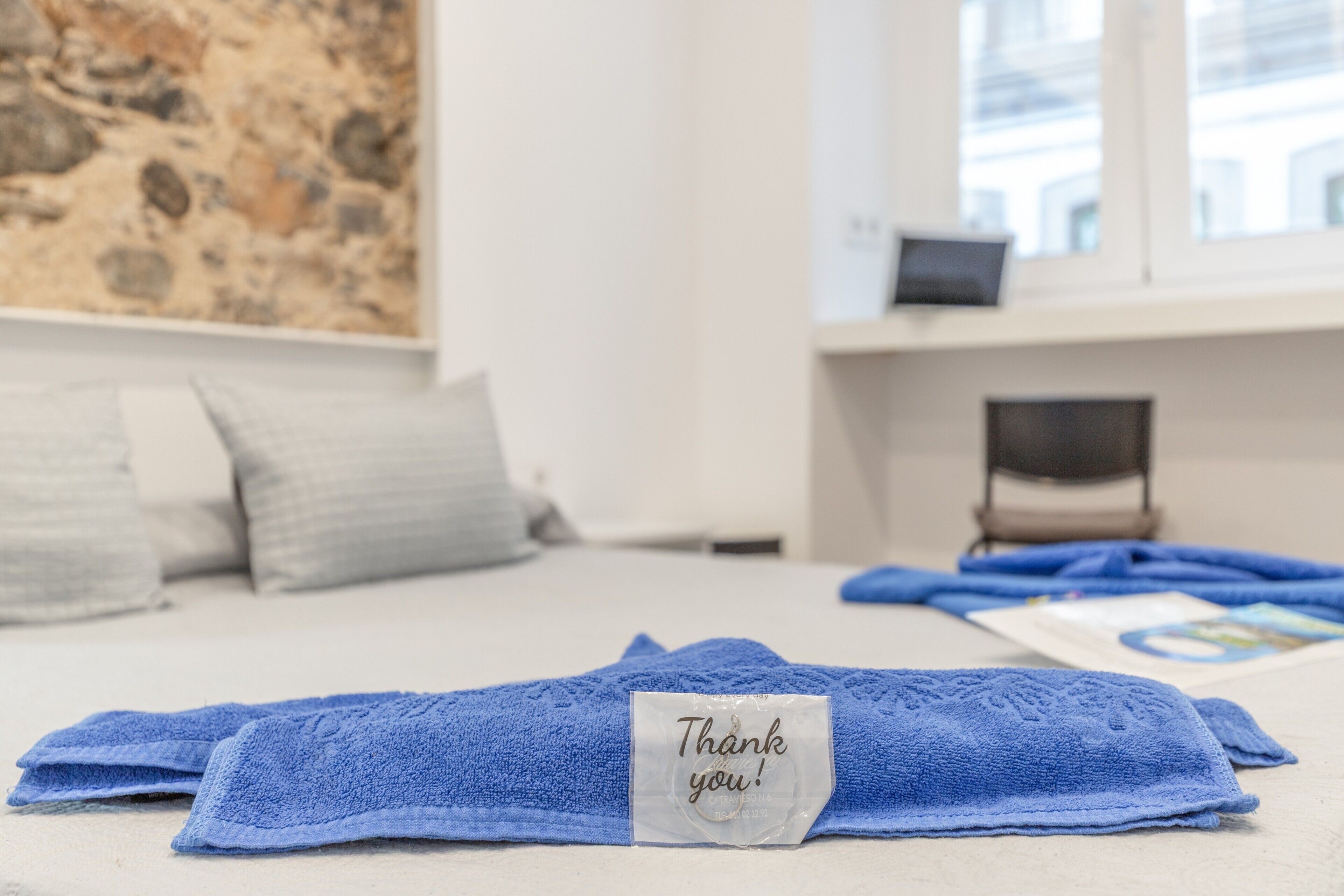 A neatly made bed with blue towels and a thank you note, a window with a view of buildings, and a painting on the wall.
