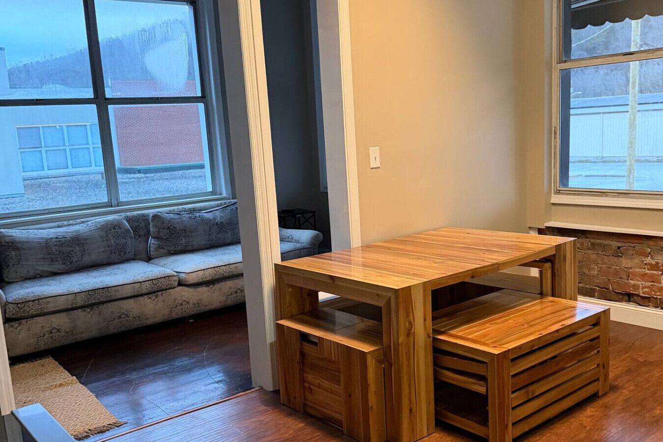 A corner room with a wooden table, a sofa, and a view through a window.