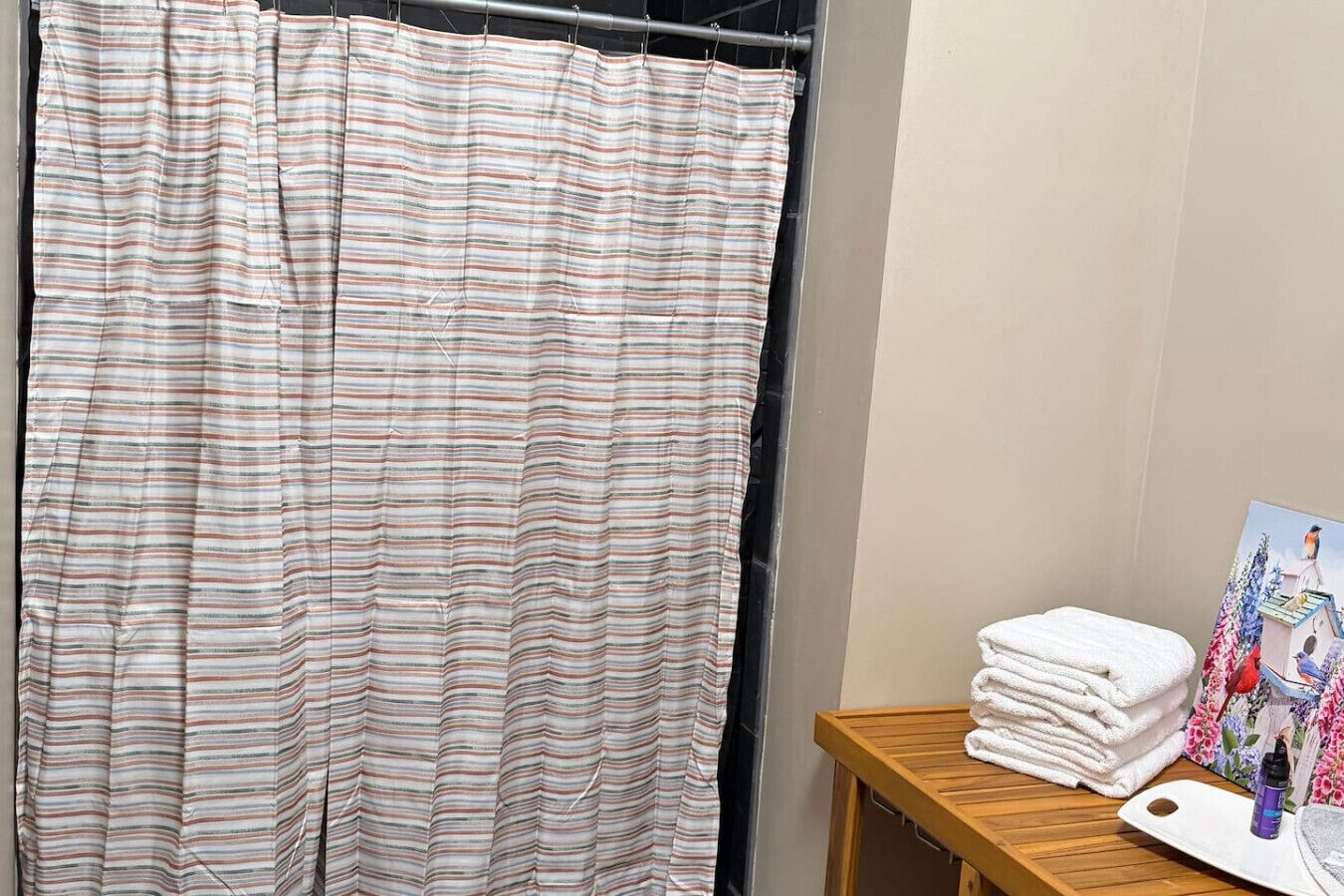 Une salle de bain avec un rideau de douche à rayures, un tabouret en bois sur lequel sont posées des serviettes et un tableau accroché au mur.