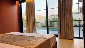 Habitación de hotel con cama, ventanas amplias y vistas a una piscina y vegetación al exterior.