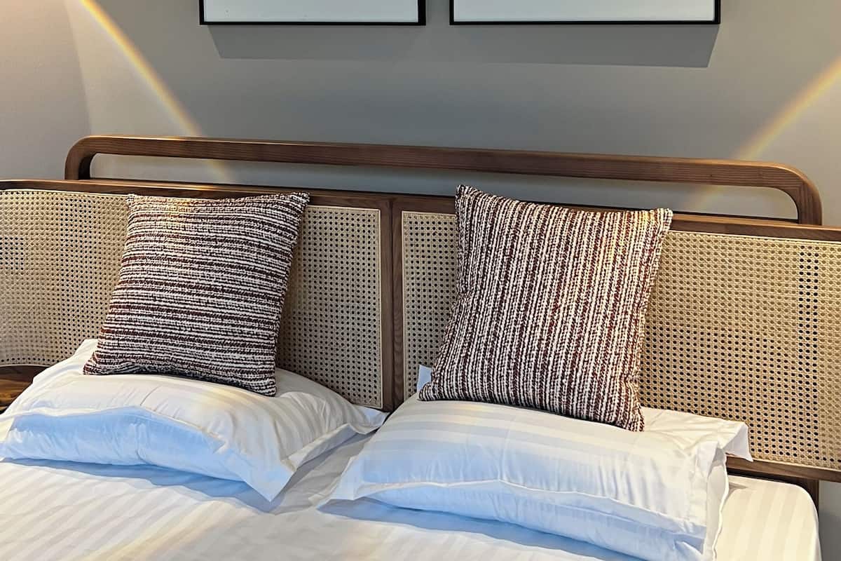 Een net opgemaakt bed met witte lakens en twee kussens met een patroon, een houten hoofdbord, en twee ingelijste abstracte kunstwerken aan de muur.
