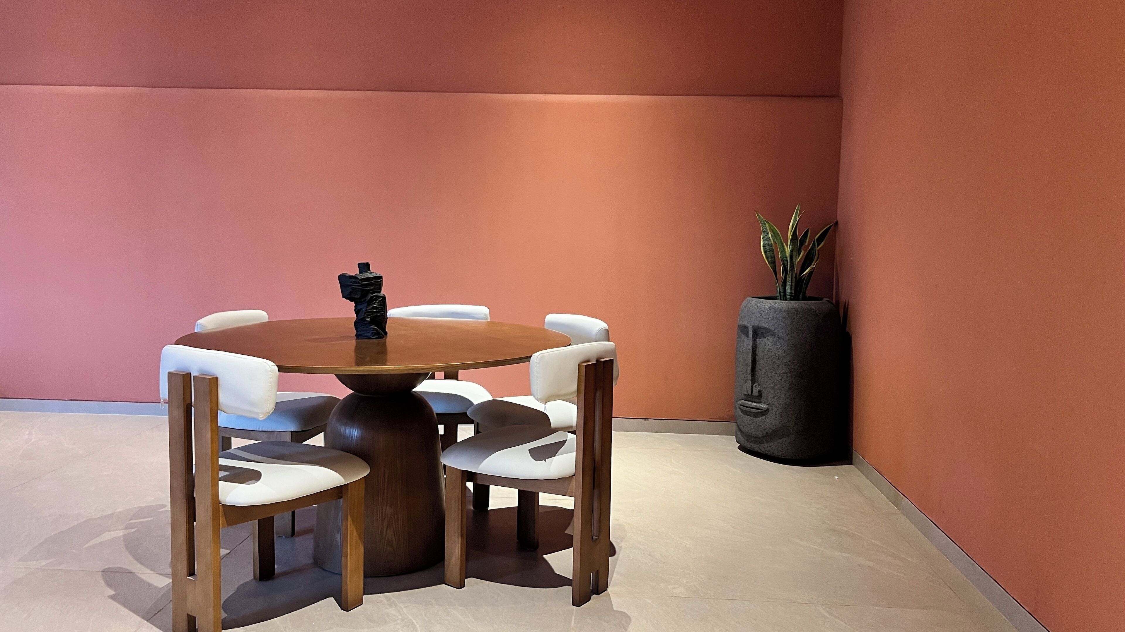 Una mesa redonda de madera con sillas blancas acolchadas en una habitación con una pared naranja lisa y una planta en maceta.