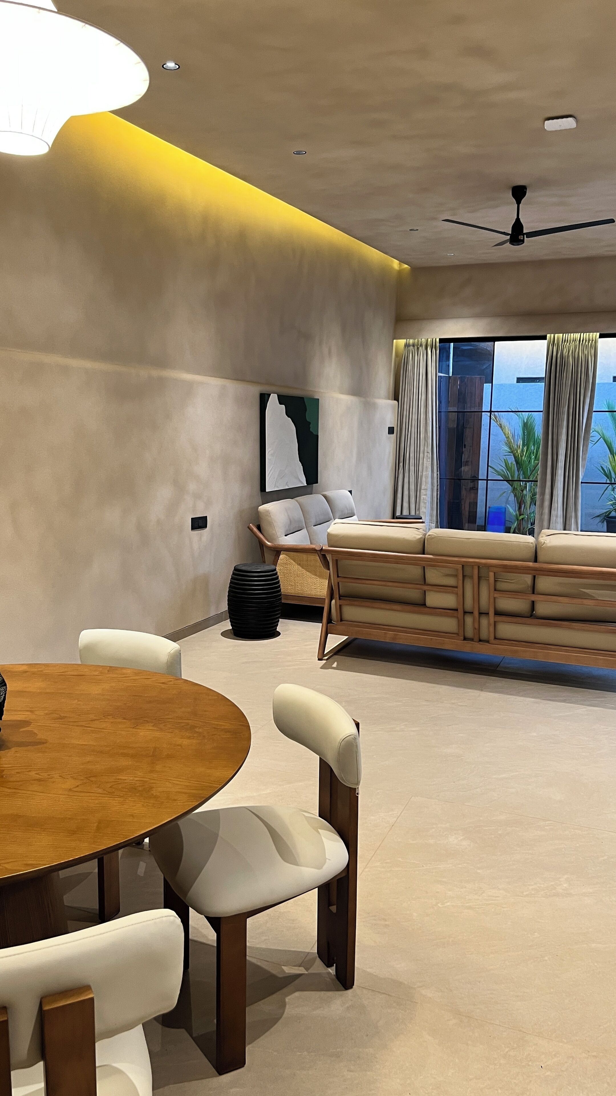 A modern living room with a round wooden table, white cushioned chairs, a sofa, and a large window with curtains.