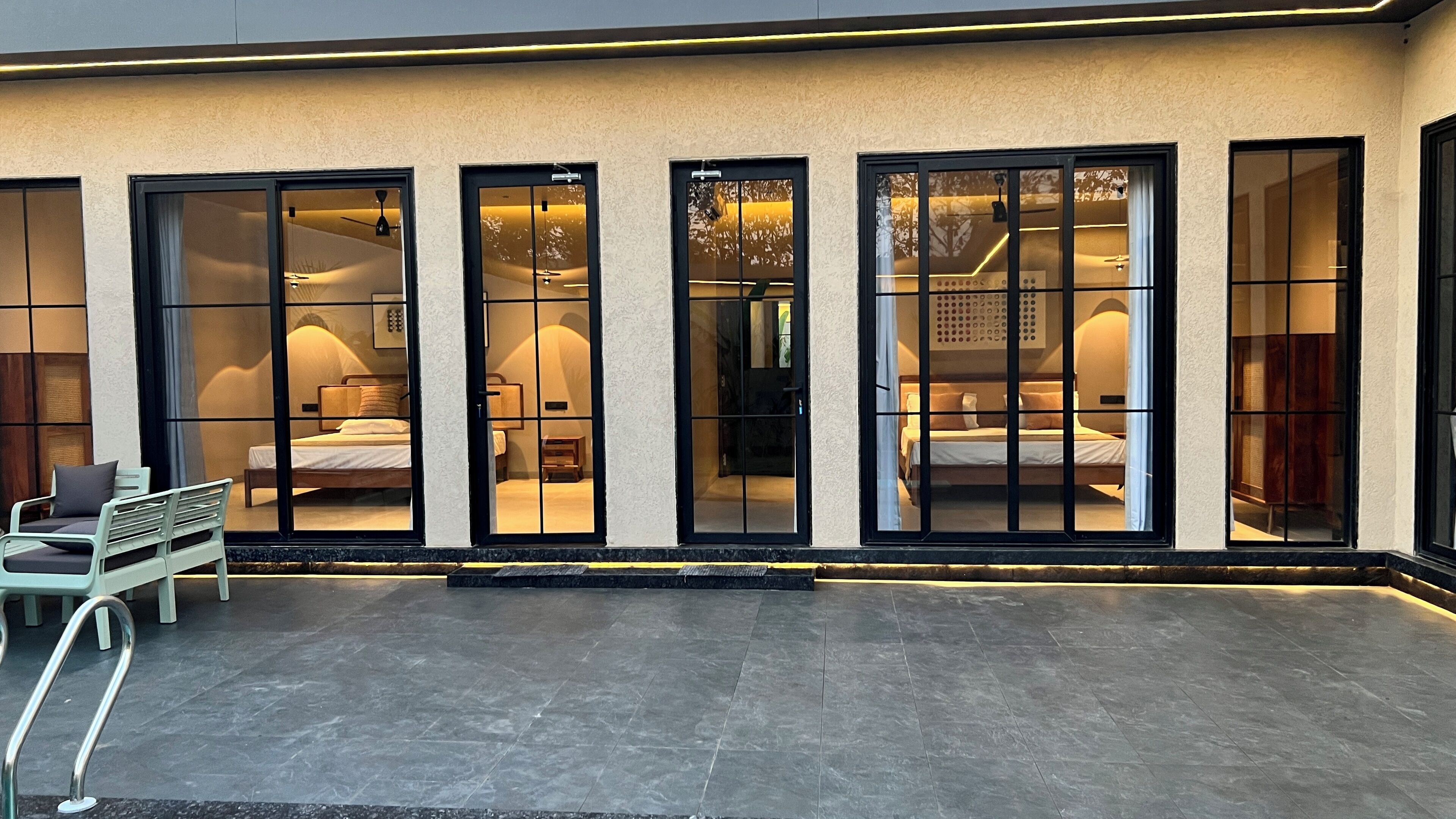 A hotel exterior with large glass doors leading to bedrooms, a tiled patio, and a metal chair.