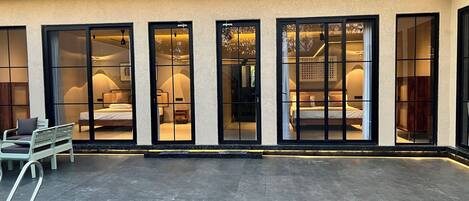 A hotel exterior with large glass doors leading to bedrooms, a tiled patio, and a metal chair.