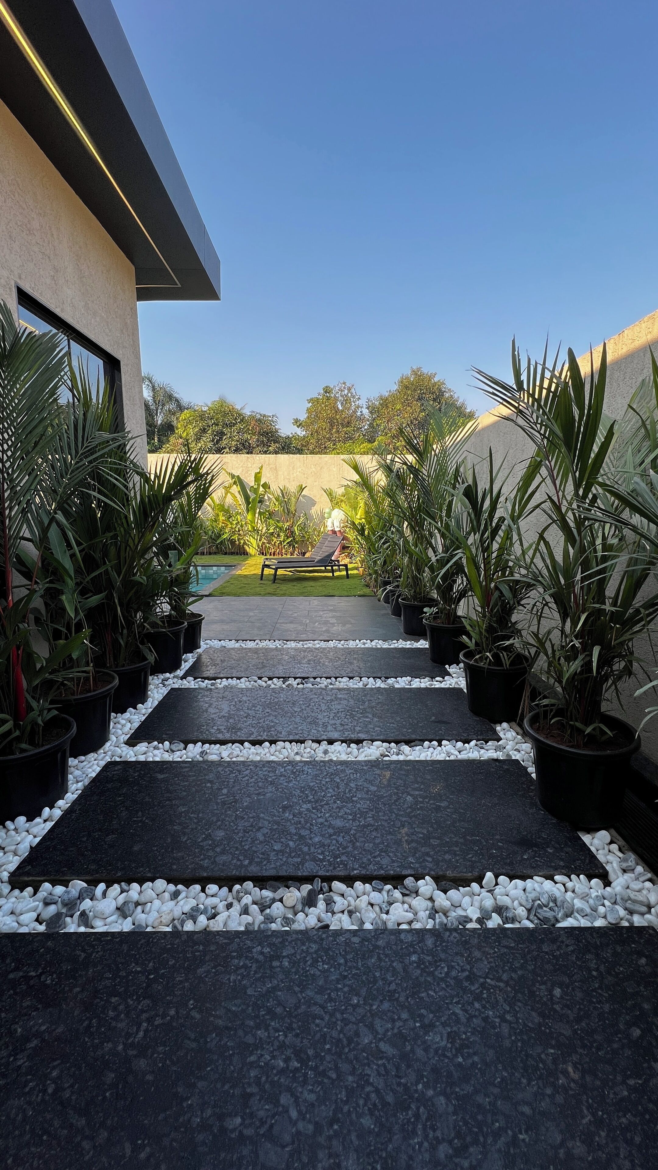 Un camino moderno al aire libre que lleva a una zona de piscina, rodeada de plantas en macetas y un cielo despejado.