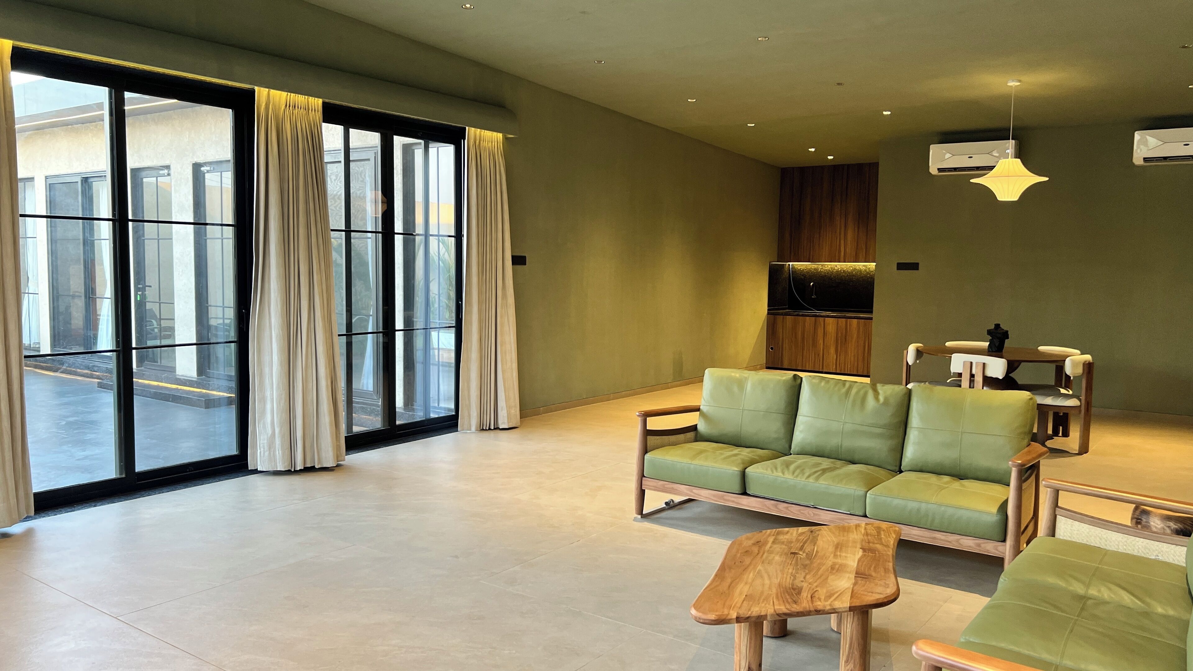 A spacious living area with a green sofa, wooden coffee table, and a dining set. Large windows with curtains provide natural light and a view of the outdoors.