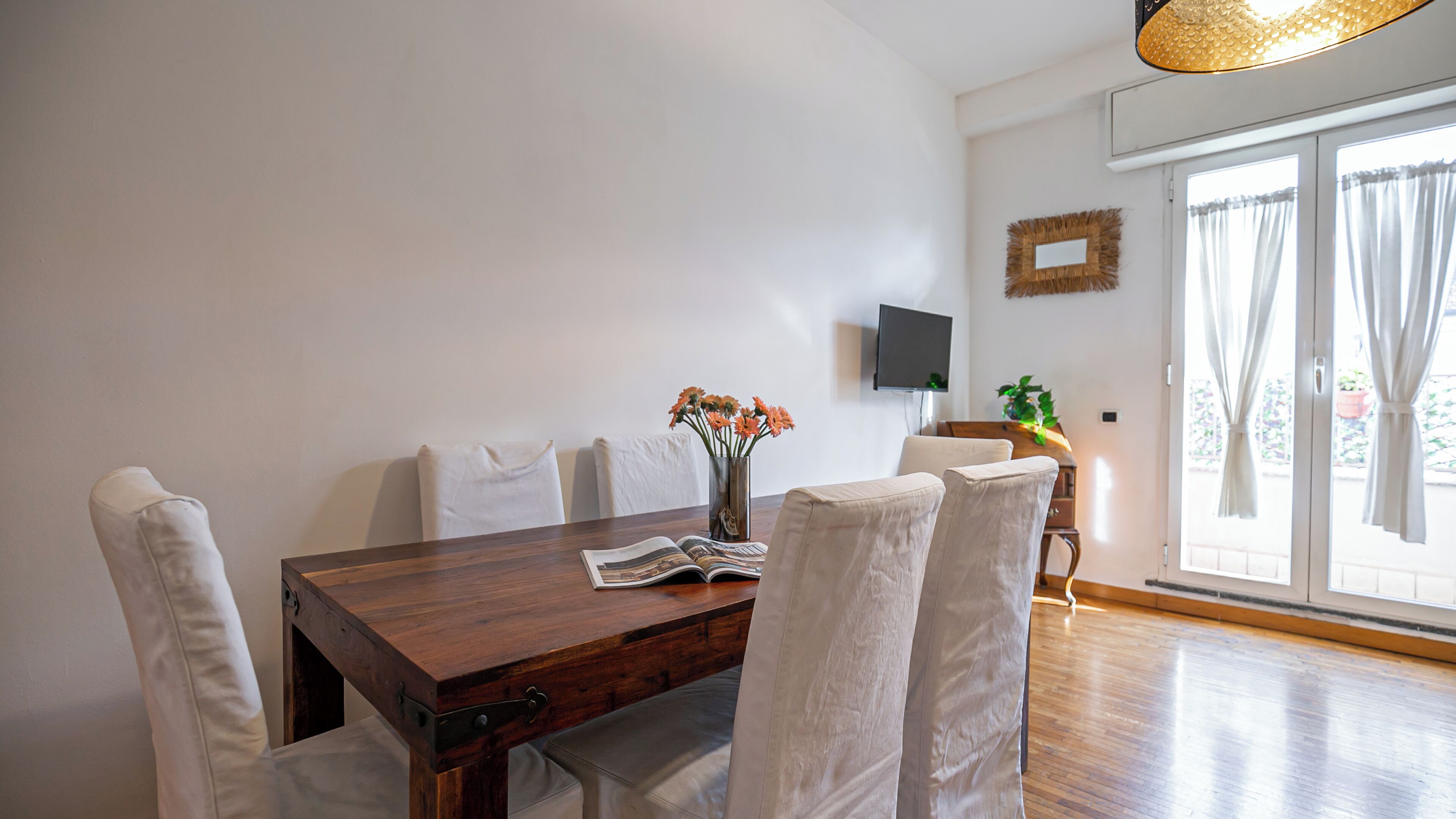 A dining area with a wooden table, white upholstered chairs, a vase with flowers, and a television on the wall.