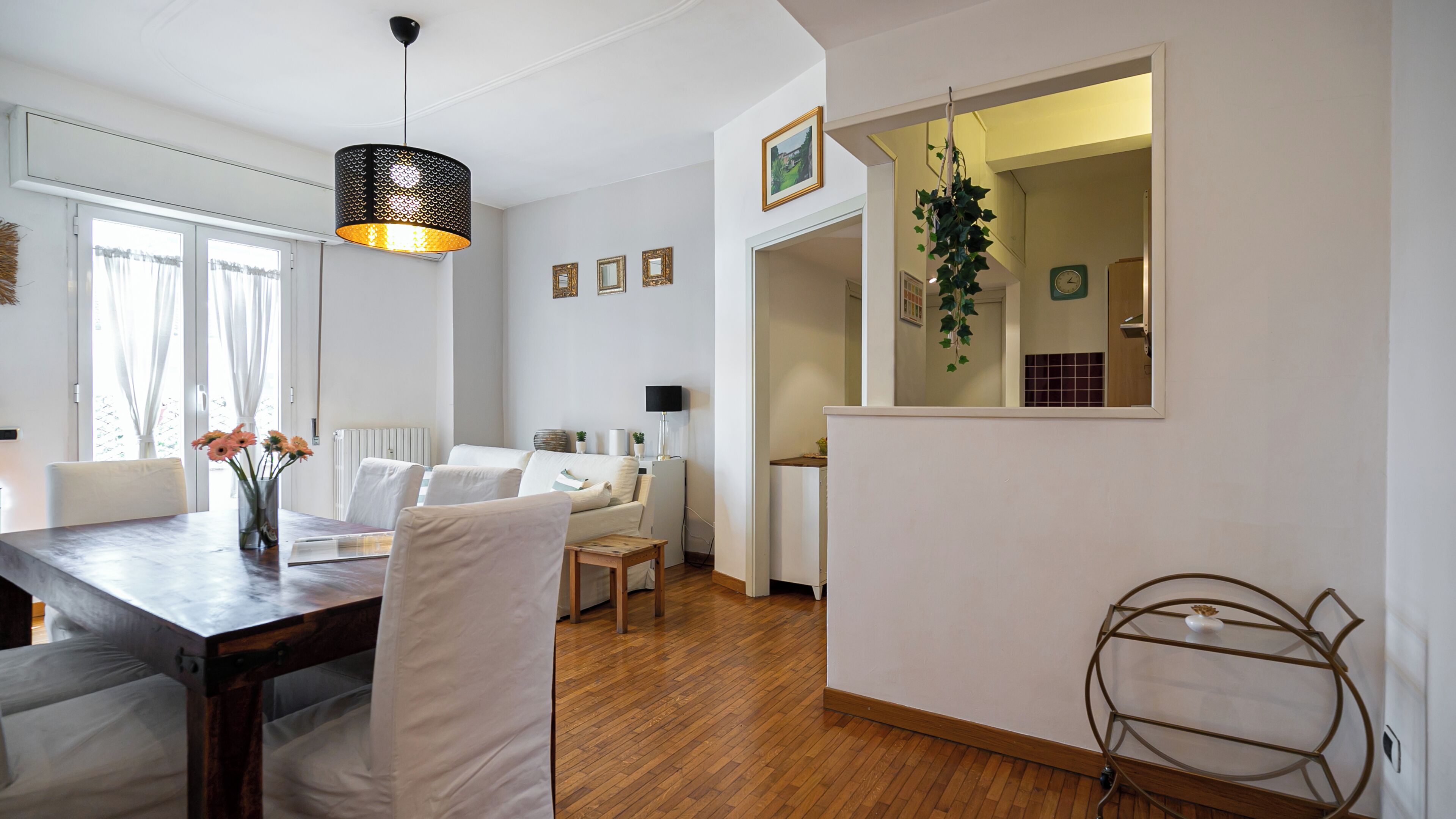 A modern living room with a dining area, white walls, wooden flooring, and a hanging plant.