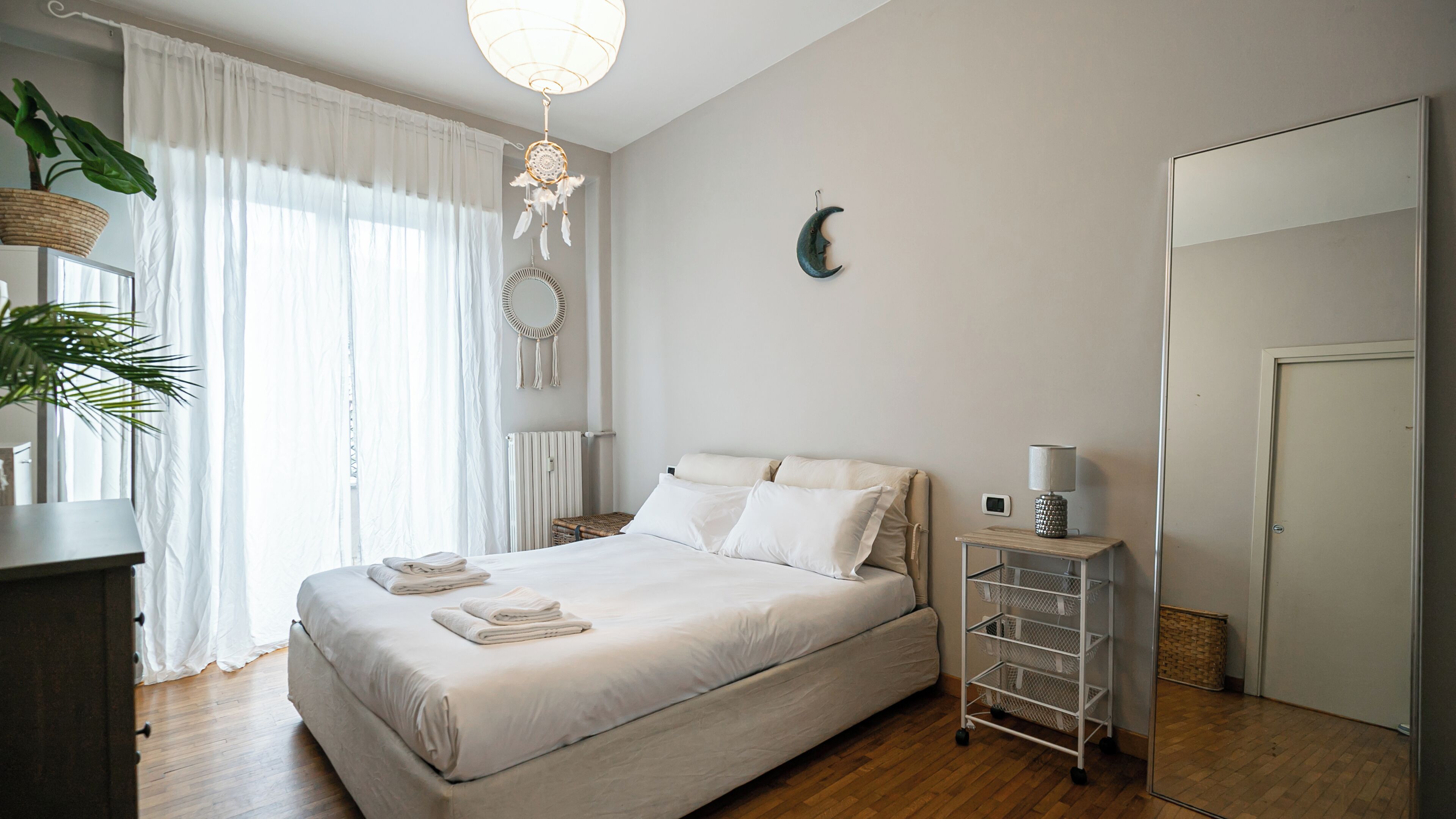 A neatly made bed with pillows and folded towels, a bedside table with a lamp, a wall-mounted dreamcatcher, and a mirror.