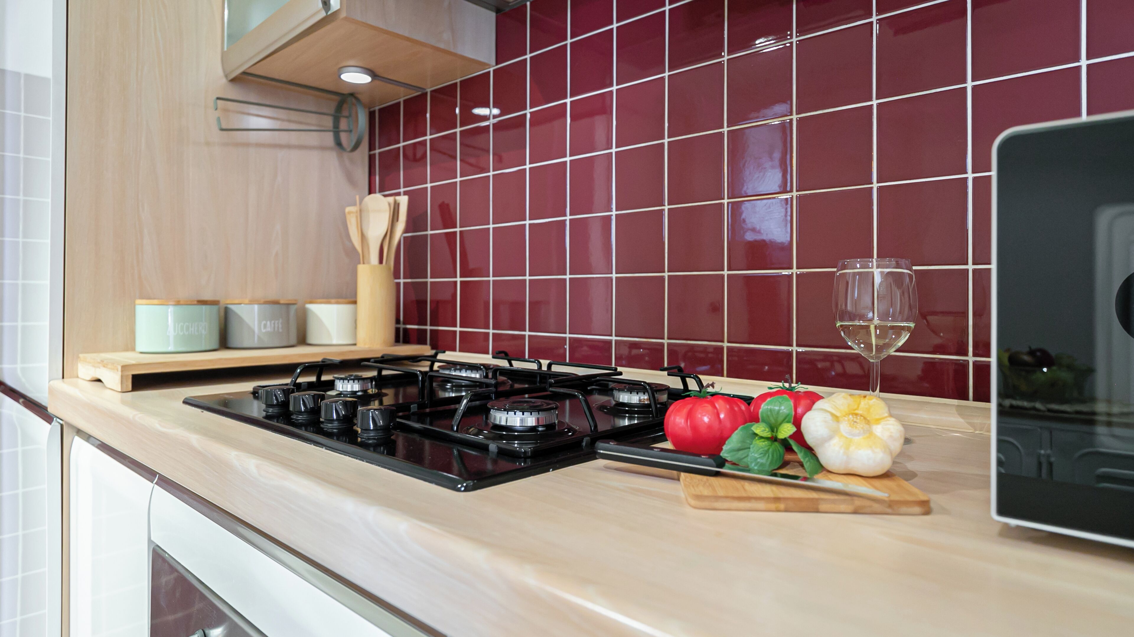 Een keuken met een gasfornuis, een rode betegelde spatwand, en diverse keukengerei zoals een snijplank, groenten en een wijnglas.