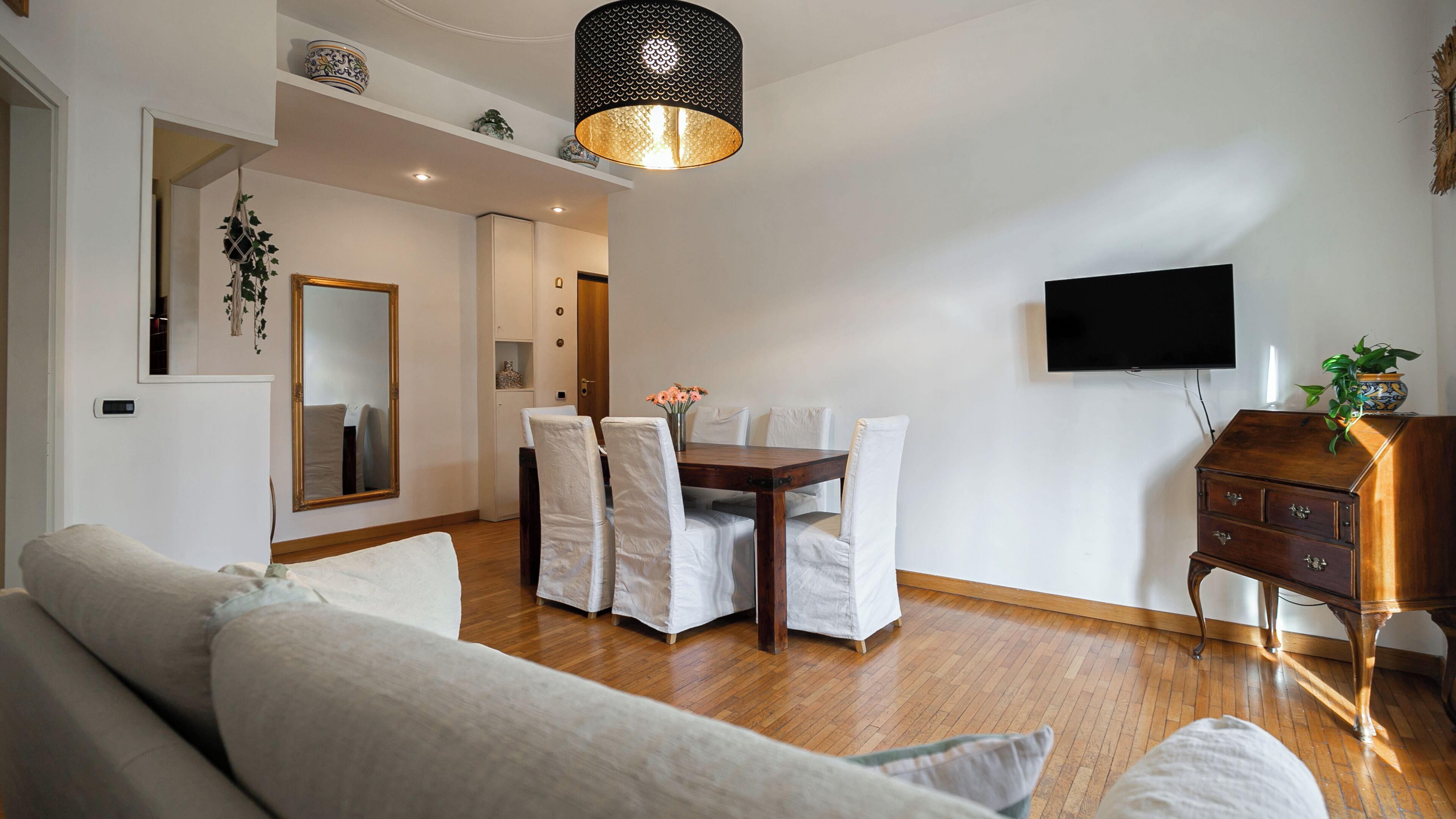 A modern living room with a wooden dining table, white upholstered chairs, a sofa, a TV mounted on the wall, and a decorative lamp.