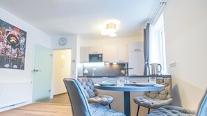 Ruokailutilassa on pyöreä pöytä ja neljä pehmustettua tuolia. Taustalla näkyy keittiö ja oikealla puolella on suuri ikkuna.