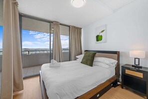 Ein Hotelzimmer mit einem großen Bett, einem Nachttisch, einer Lampe und Blick auf die Stadt durch ein großes Fenster.