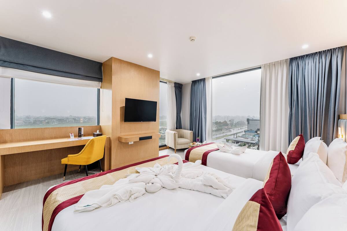Une chambre d’hôtel équipée d’un bureau, d’une chaise, de deux lits, d’une télévision et de grandes fenêtres avec des rideaux.