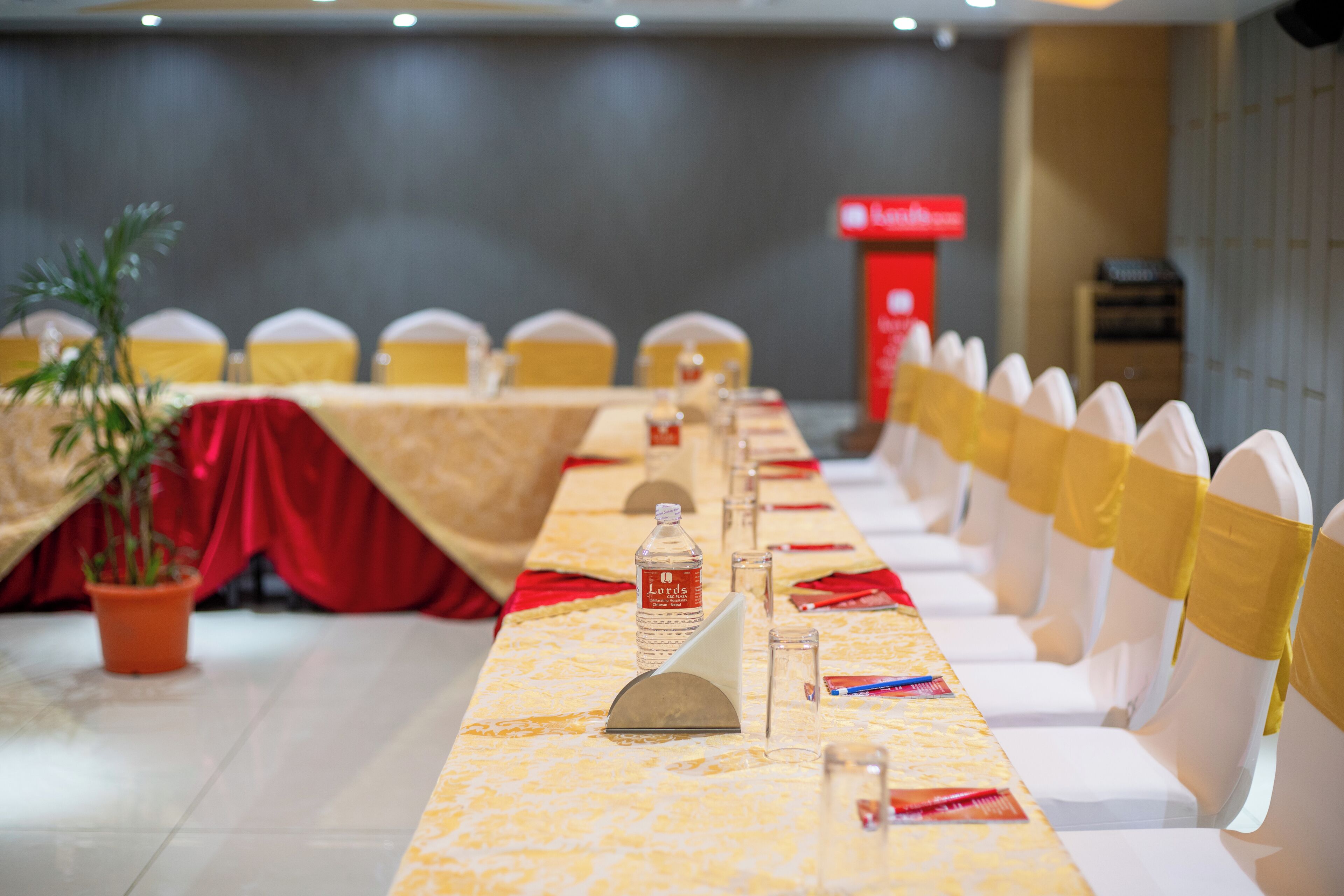 Una sala de conferencias con una mesa larga, sillas con cintas amarillas, una botella de agua y una cortina roja.