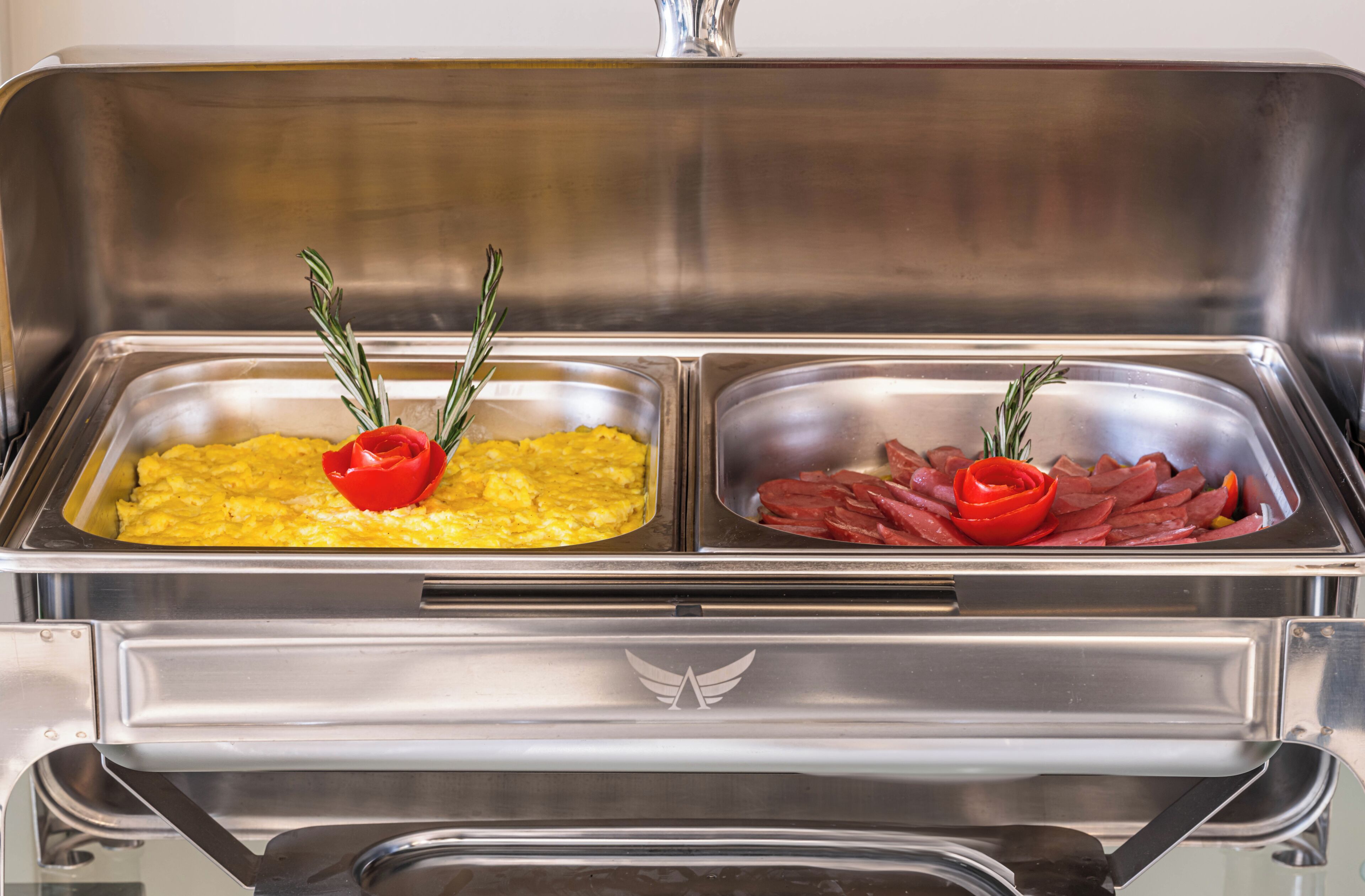 En buffet med to rustfrie stålfade, hvoraf det ene indeholder røræg pyntet med rød peberfrugt og rosmarin, og det andet indeholder skiveskåret kød og en rød peberfrugt.