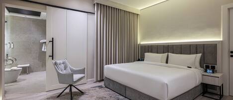 Un dormitorio moderno con una cama grande, una mesita de noche con un reloj, una silla y un baño visible a través de una puerta abierta.
