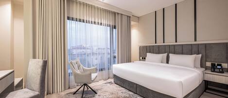 A modern hotel room with a large bed, a desk, a chair, and a view of the city through large windows.
