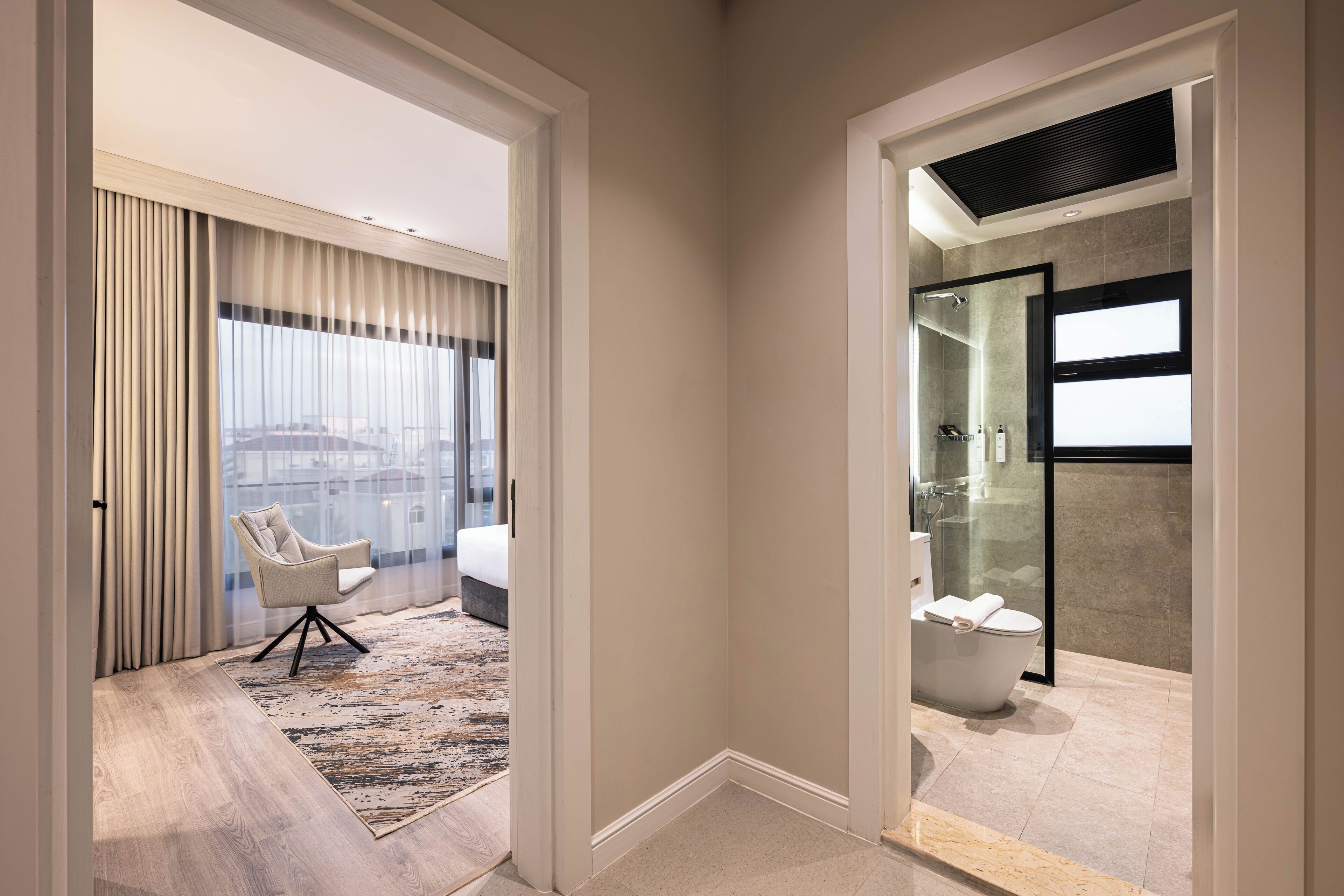 A modern hotel room with a large window, a sitting area with a chair, a bathroom with a bathtub, and a shower area.