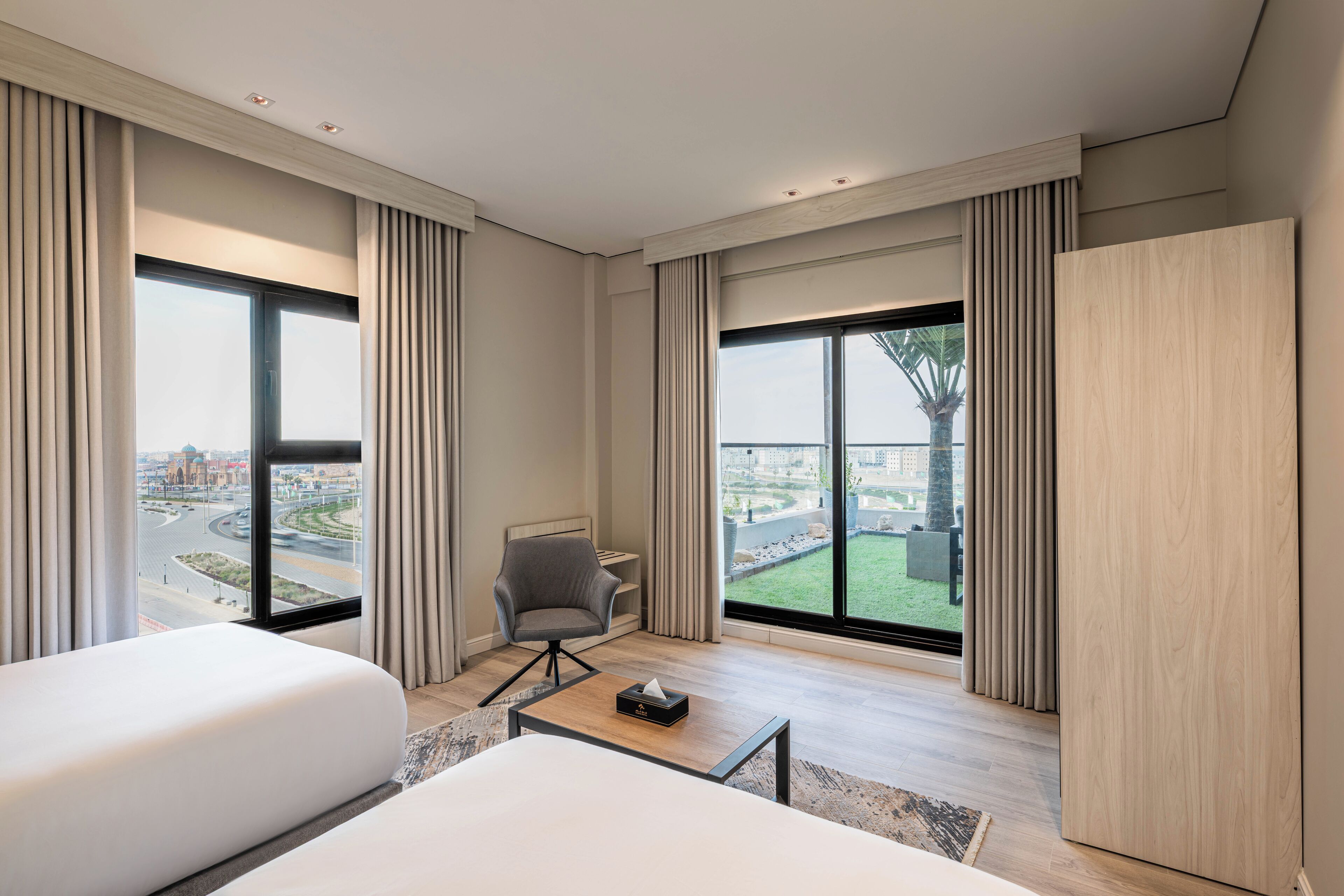 Ein modernes Hotelzimmer mit einem großen Bett, einem Schreibtisch und einem Stuhl. Große Fenster mit Vorhängen, Blick auf die Stadt und ein Balkon mit Baum.