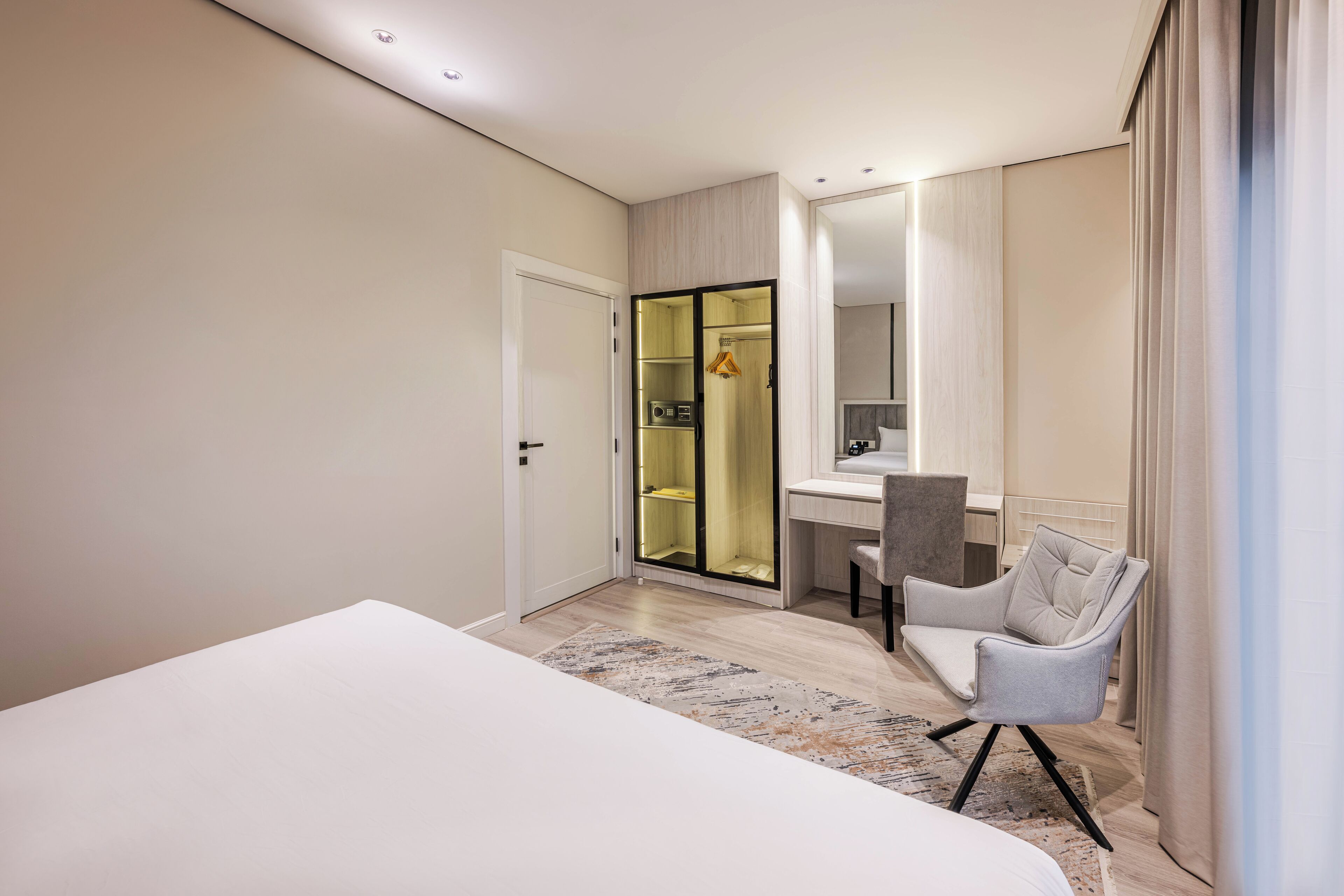 A modern hotel room with a large bed, a desk with a chair, a wardrobe, and a bathroom with a shower.