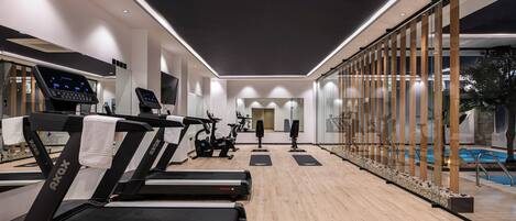 A modern gym with treadmills, exercise bikes, and a pool area visible through glass walls.