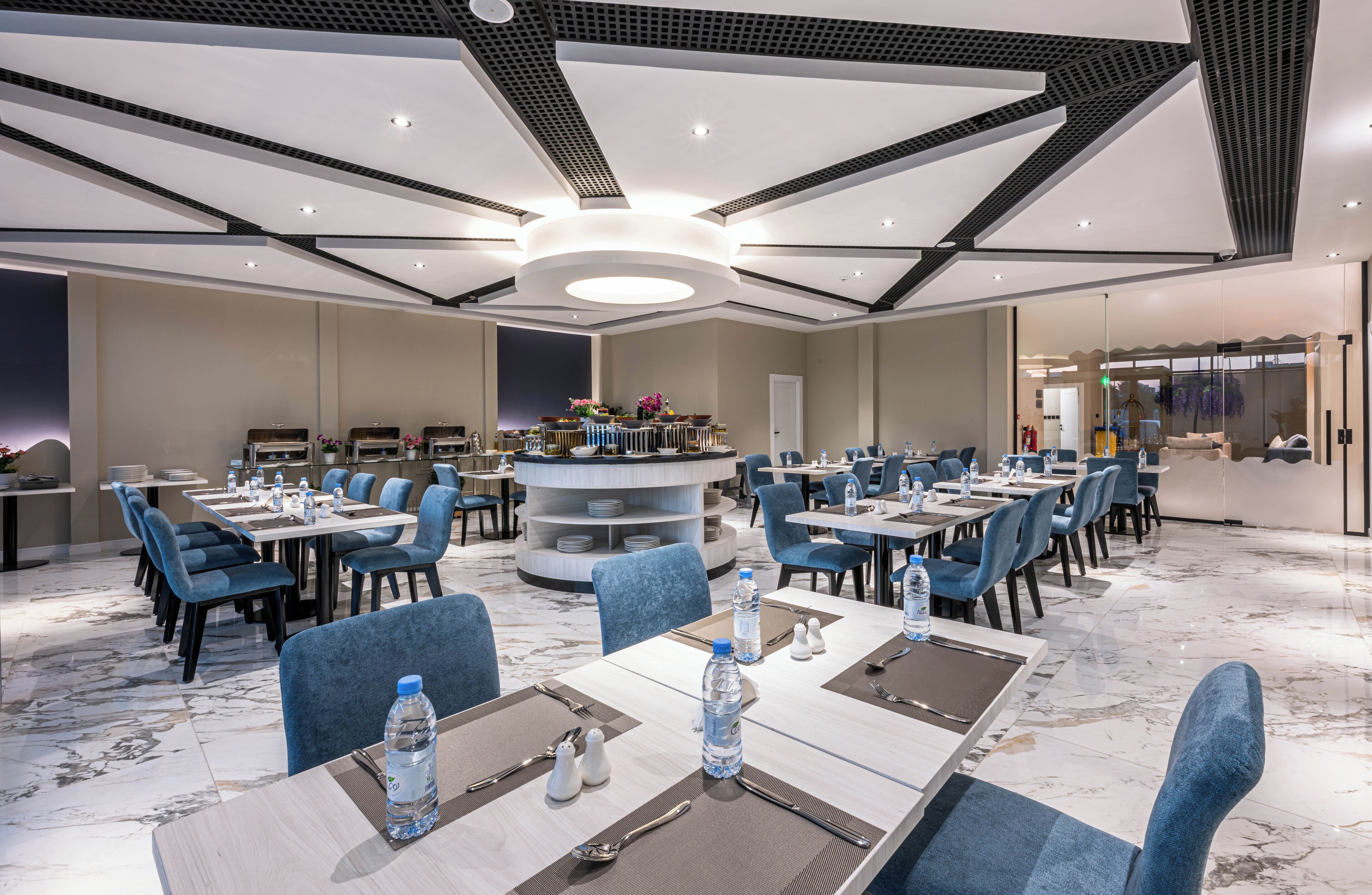 A modern hotel dining area with round tables, blue chairs, and a central water feature.