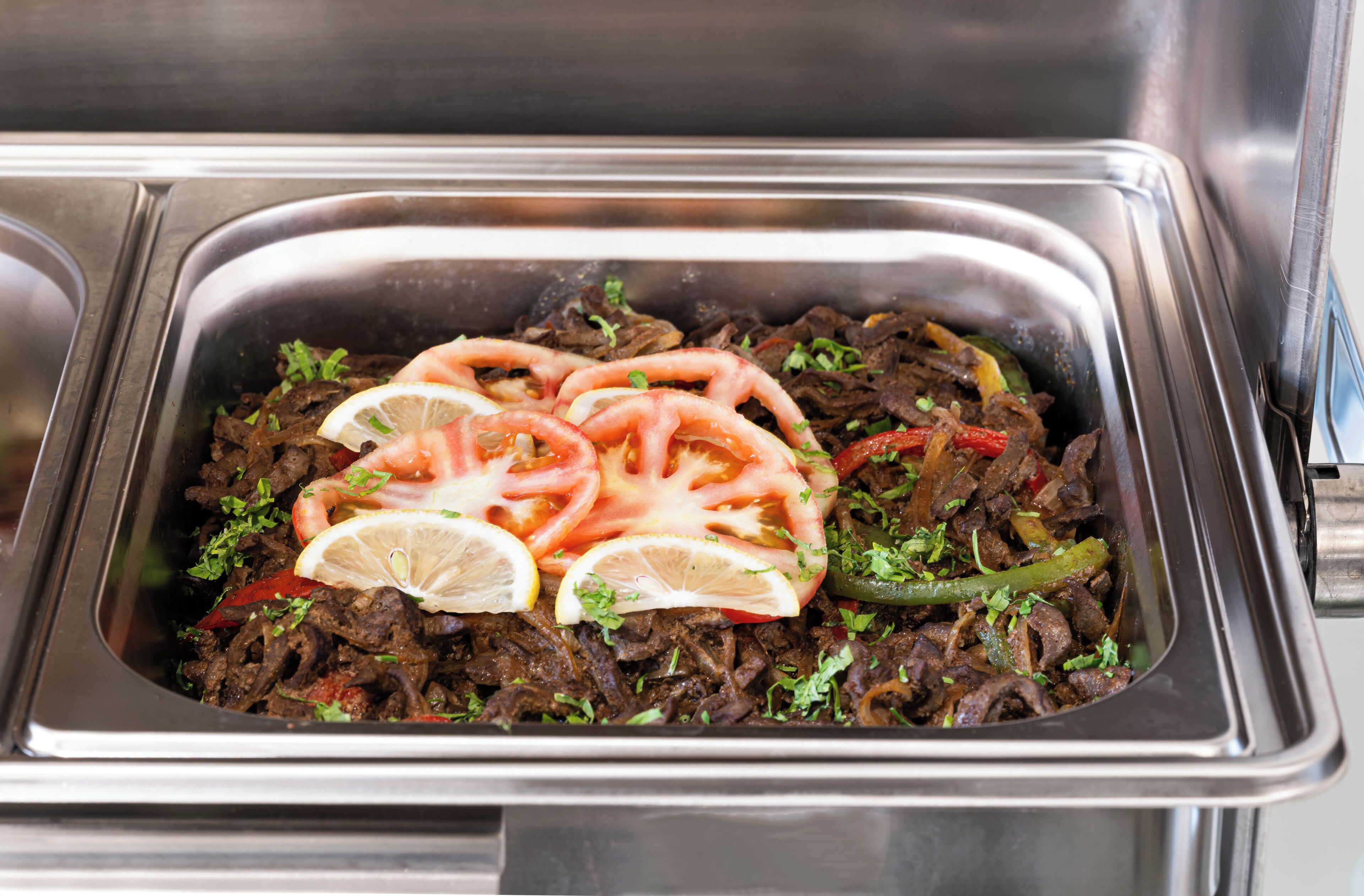 A stainless steel serving dish with a meal consisting of shredded meat, tomatoes, and green peppers, garnished with lemon slices and herbs.