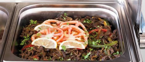 A stainless steel serving dish with a meal consisting of shredded meat, tomatoes, and green peppers, garnished with lemon slices and herbs.
