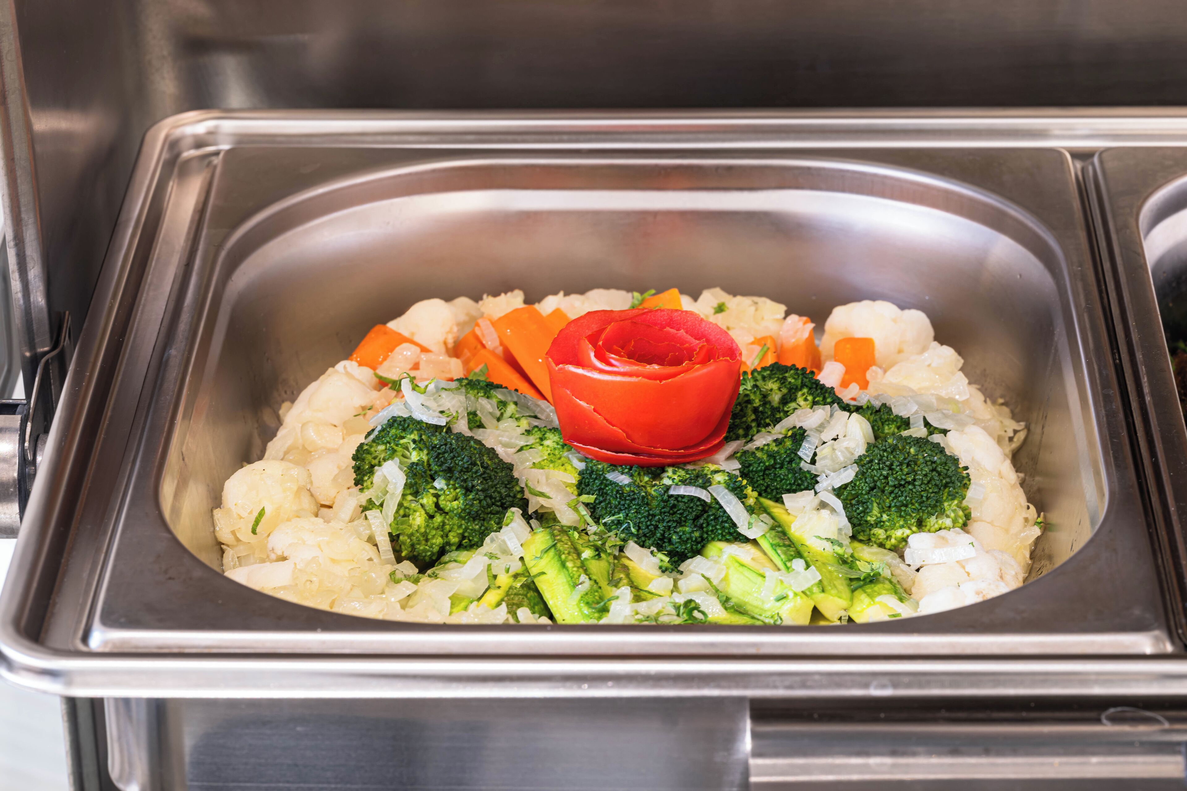 A stainless steel food serving dish with a variety of vegetables including broccoli, cauliflower, carrots, and a decorative red tomato.