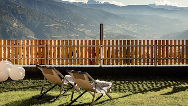 Blick auf eine gebirgige Landschaft von einer Holzdielen-Terrasse mit zwei Liegestühlen und weißen Ballons.