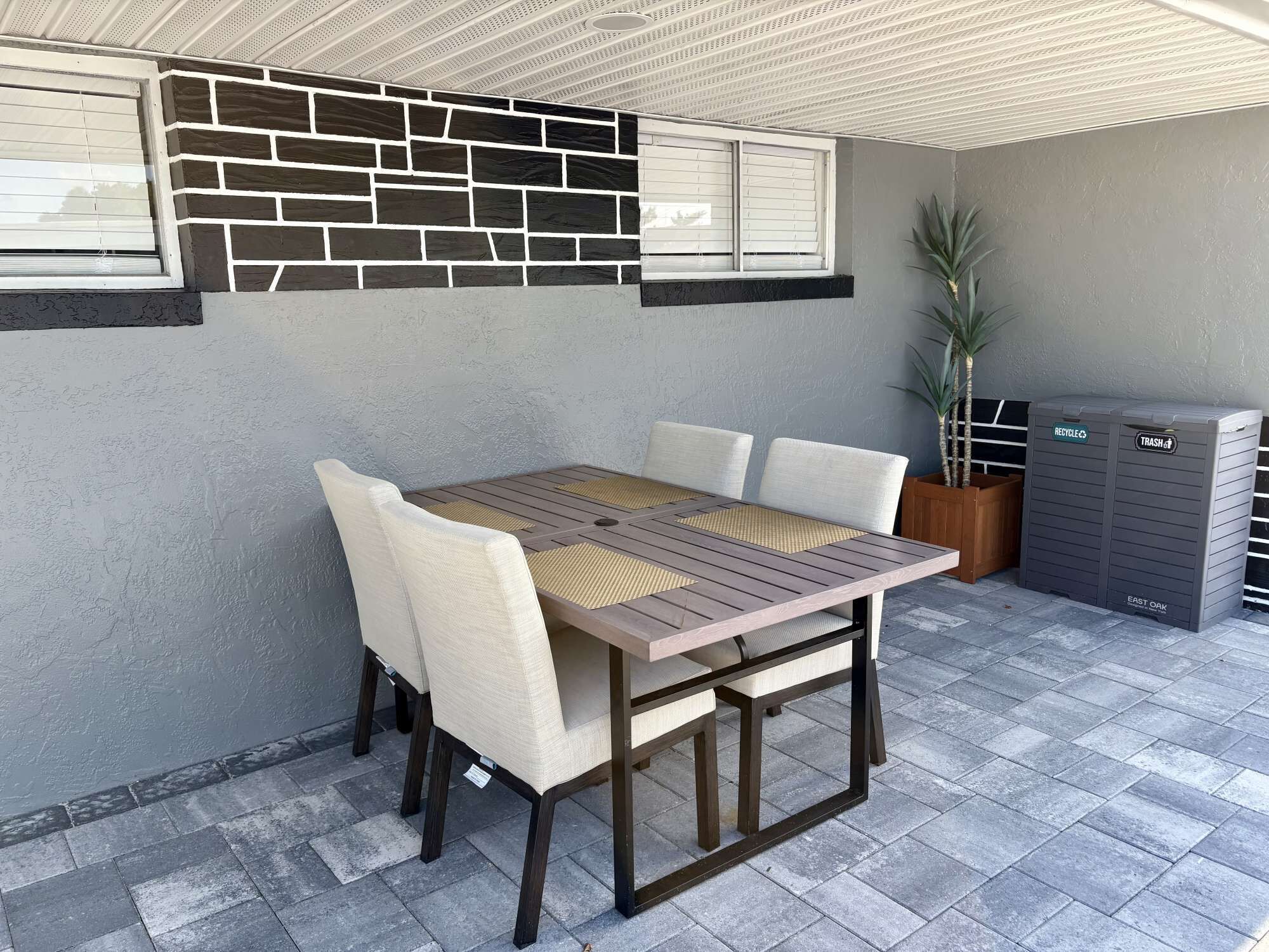 A patio with a wooden table and chairs, a potted plant, and a trash bin.