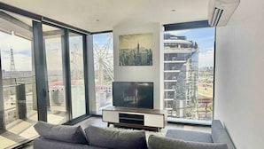 A modern living room with a large gray sofa, a flat-screen TV, and a cityscape view through large windows. - Cosy 2 Bedroom Apartment With Pool, gym and Sauna (Melbourne)