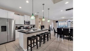 A modern kitchen with white cabinets, a stainless steel refrigerator, a microwave, and a large island with bar stools. - Spacious 7 BR Villa with Pool, Spa, Themed Rooms (Kissimmee)