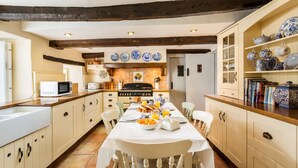 A kitchen with a long table set for breakfast, wooden beams on the ceiling, and a variety of dishes on shelves.