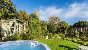 A backyard with a hot tub, a wooden deck, and a garden with various plants and a white statue.
