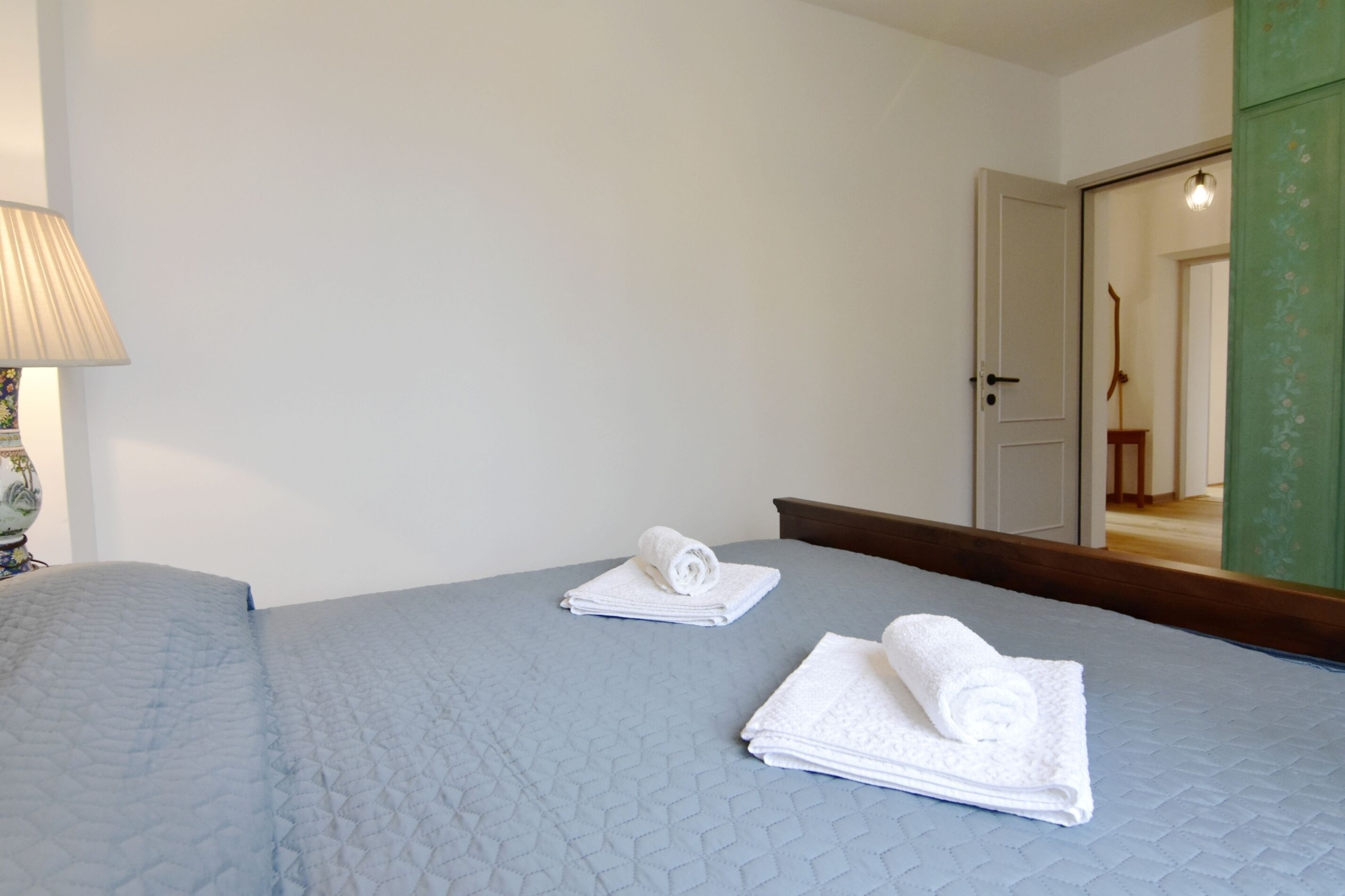 Une chambre d’hôtel avec un lit, deux serviettes enroulées, une lampe et un miroir qui reflète une porte et une chaise.