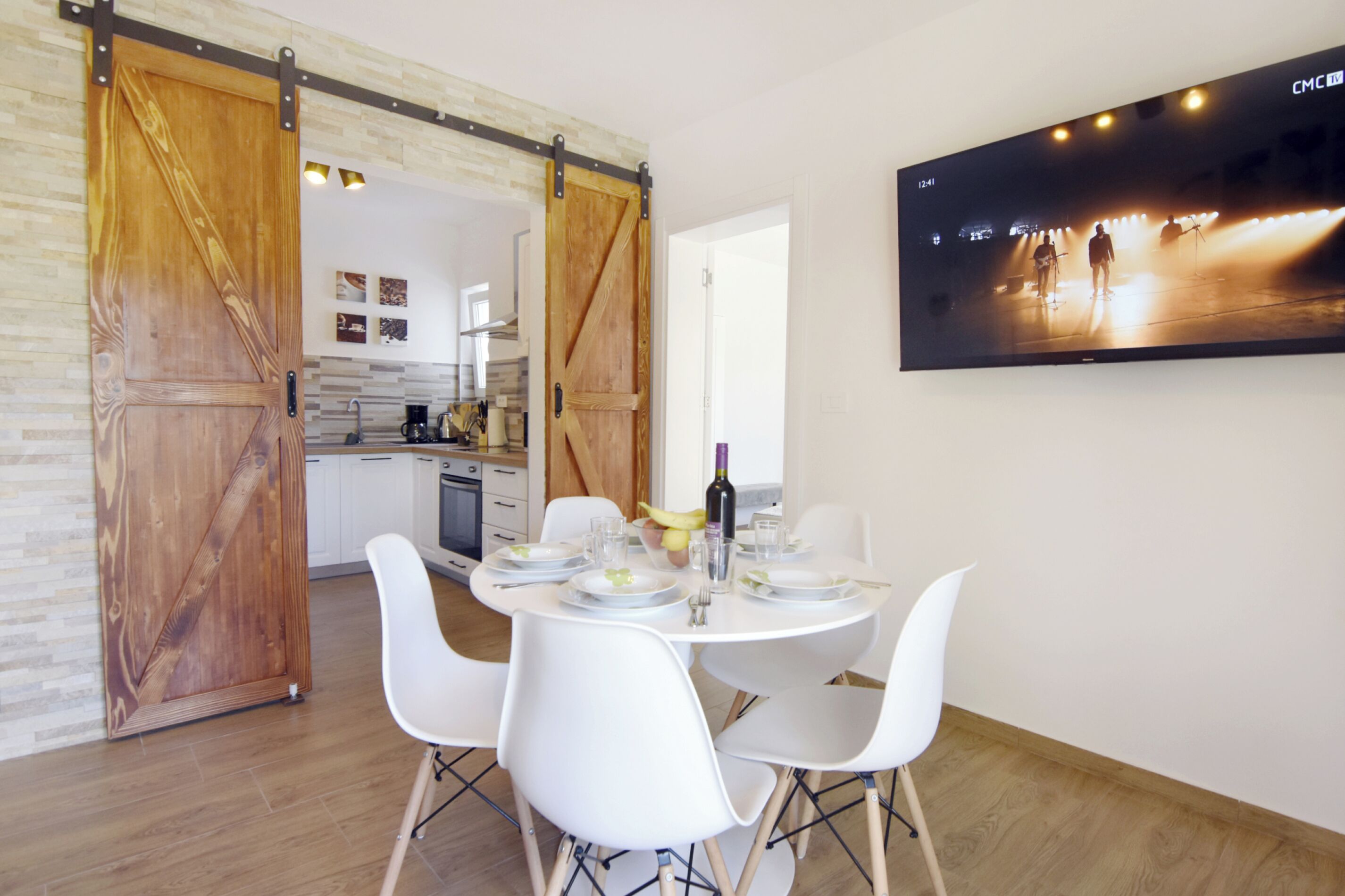 Zona pranzo con tavolo bianco imbandito per quattro, sedie bianche e un'ampia TV a schermo piatto appesa al muro che mostra una scena di concerto.