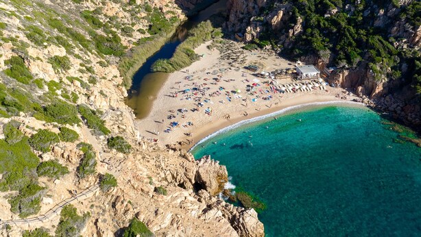 Vista aérea de una playa costera con acantilados rocosos, aguas turquesas cristalinas y una zona de arena con sombrillas y gente.