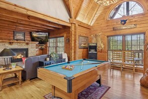 A cozy log cabin interior with a pool table, fireplace, and large windows.