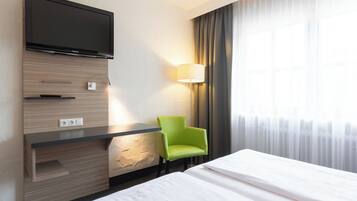 A hotel room with a flat-screen TV mounted on a wooden panel, a green chair, a bedside table, a lamp, and a bed with white linens.